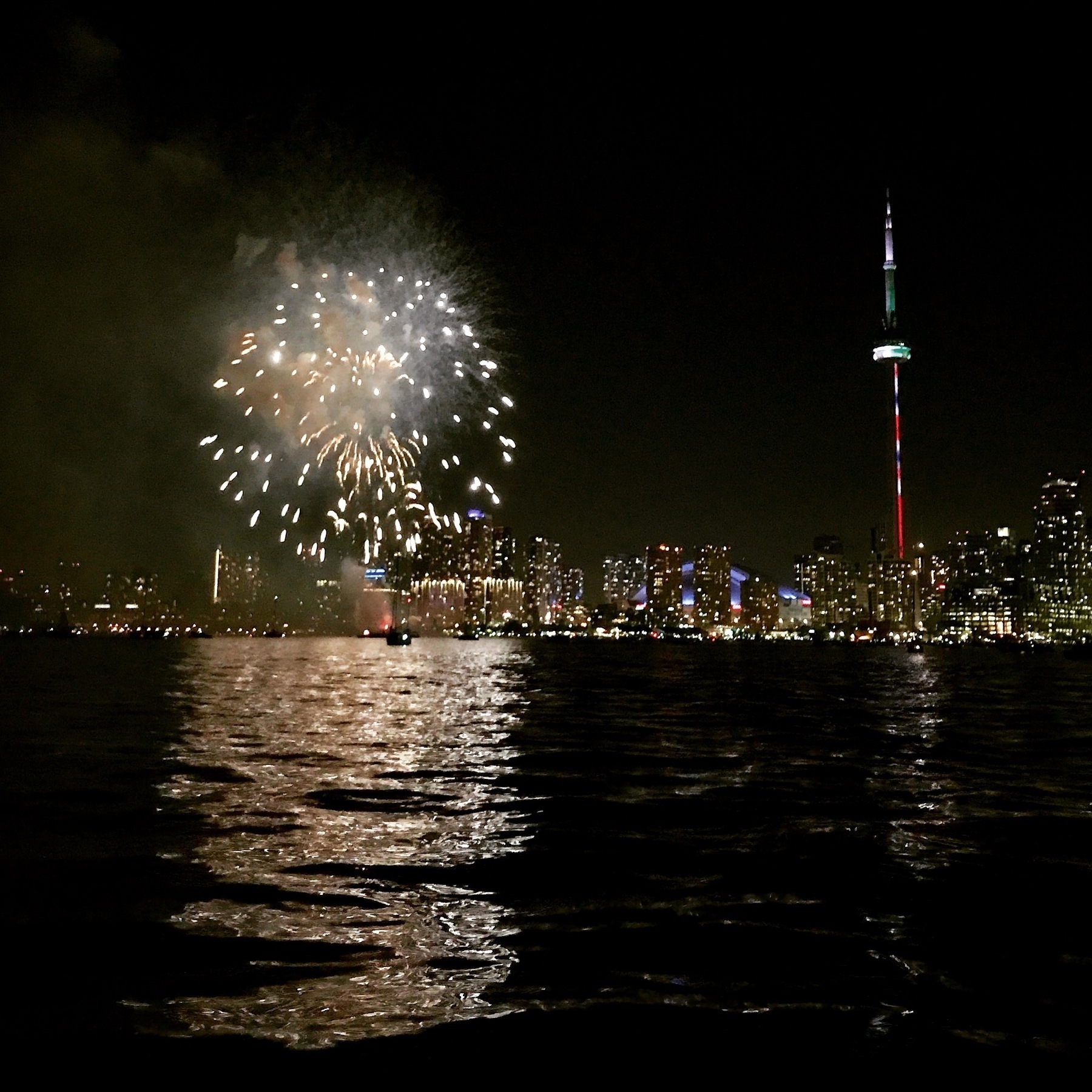 Fireworks on the left with CN Tower light up on the right against a dark sky. Taken from the lake with reflections on the water