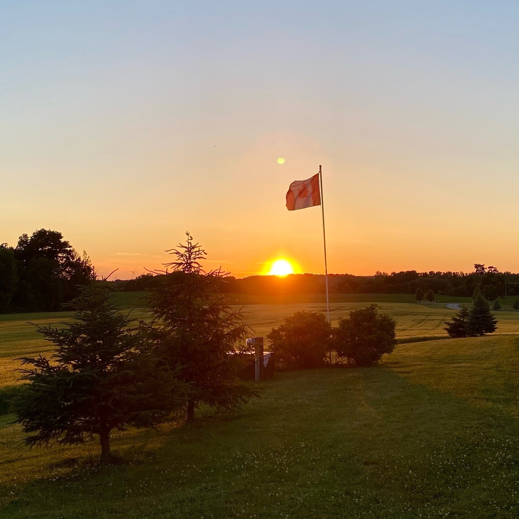 Sun setting over a field with a Canadian flag flying in the foreground