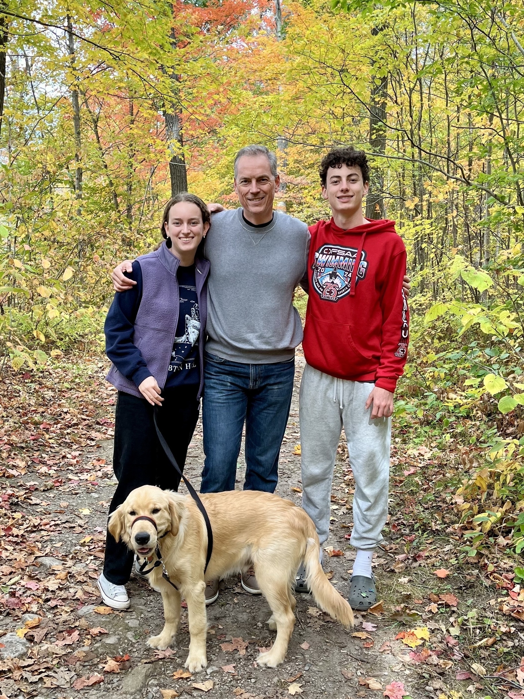 A man and two young people stand together with a golden retriever on a forest path surrounded by colorful fall foliage.