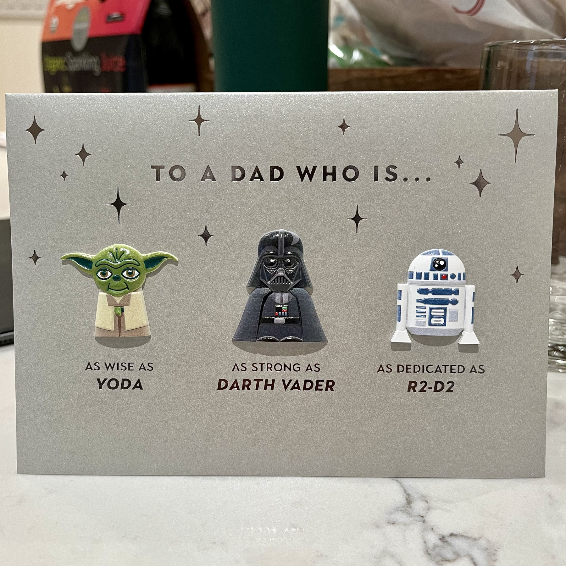 Greeting card with illustrations of Yoda, Darth Vader, and R2-D2 from Star Wars. Text reads: “TO A DAD WHO IS… AS WISE AS YODA AS STRONG AS DARTH VADER AS DEDICATED AS R2-D2”. Stars decorate background.