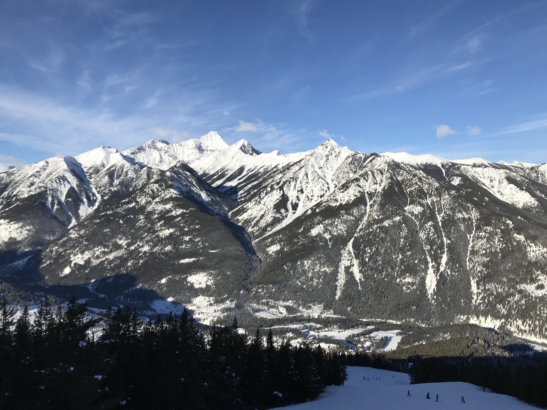 Snow-covered mountains under a blue sky, with small figures skiing on slopes in the foreground, amidst a vast, rugged alpine landscape.