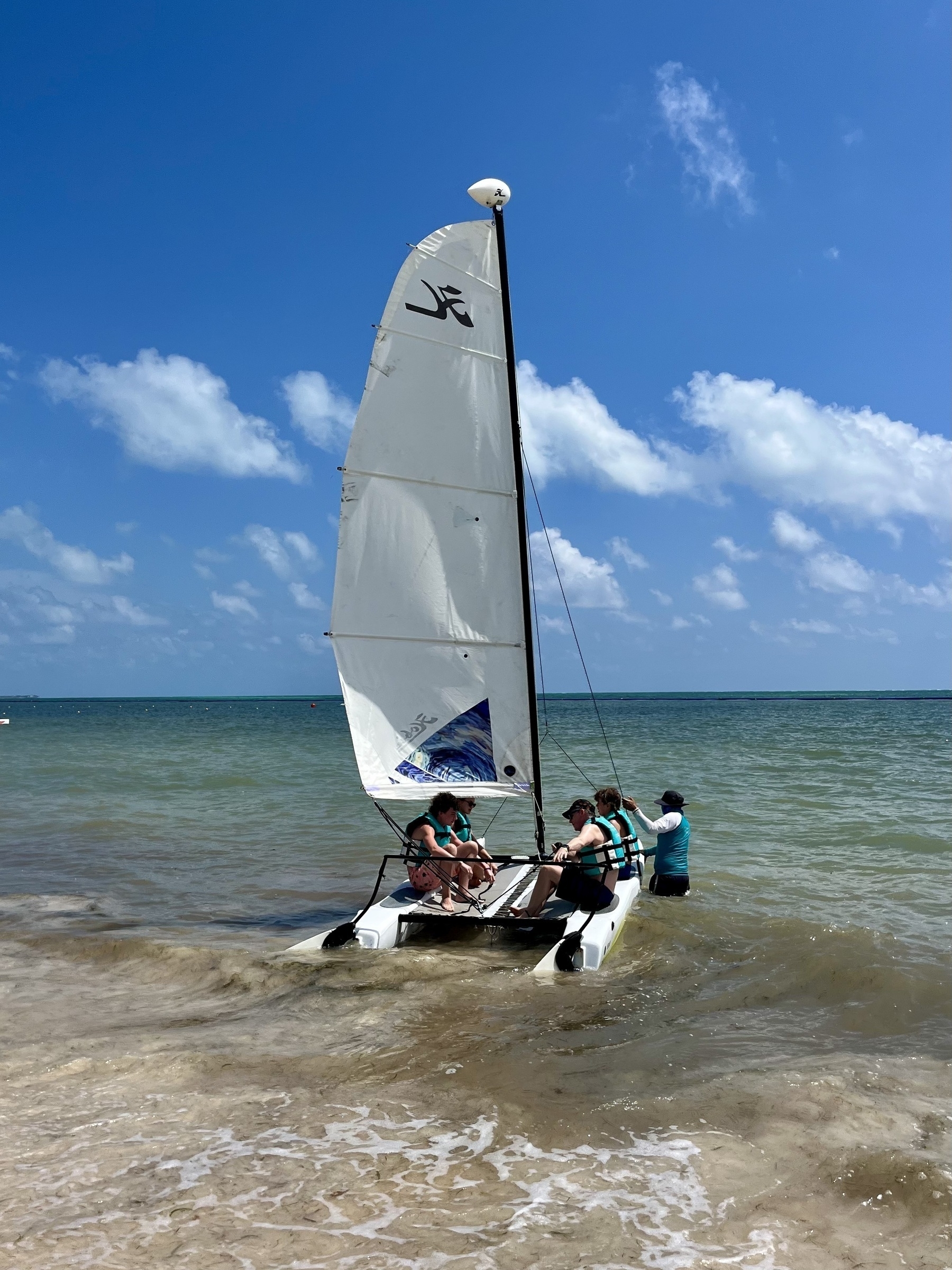 A catamaran with a raised sail is on the shore, with people preparing to sail in a sunny, beachside setting with clear skies.