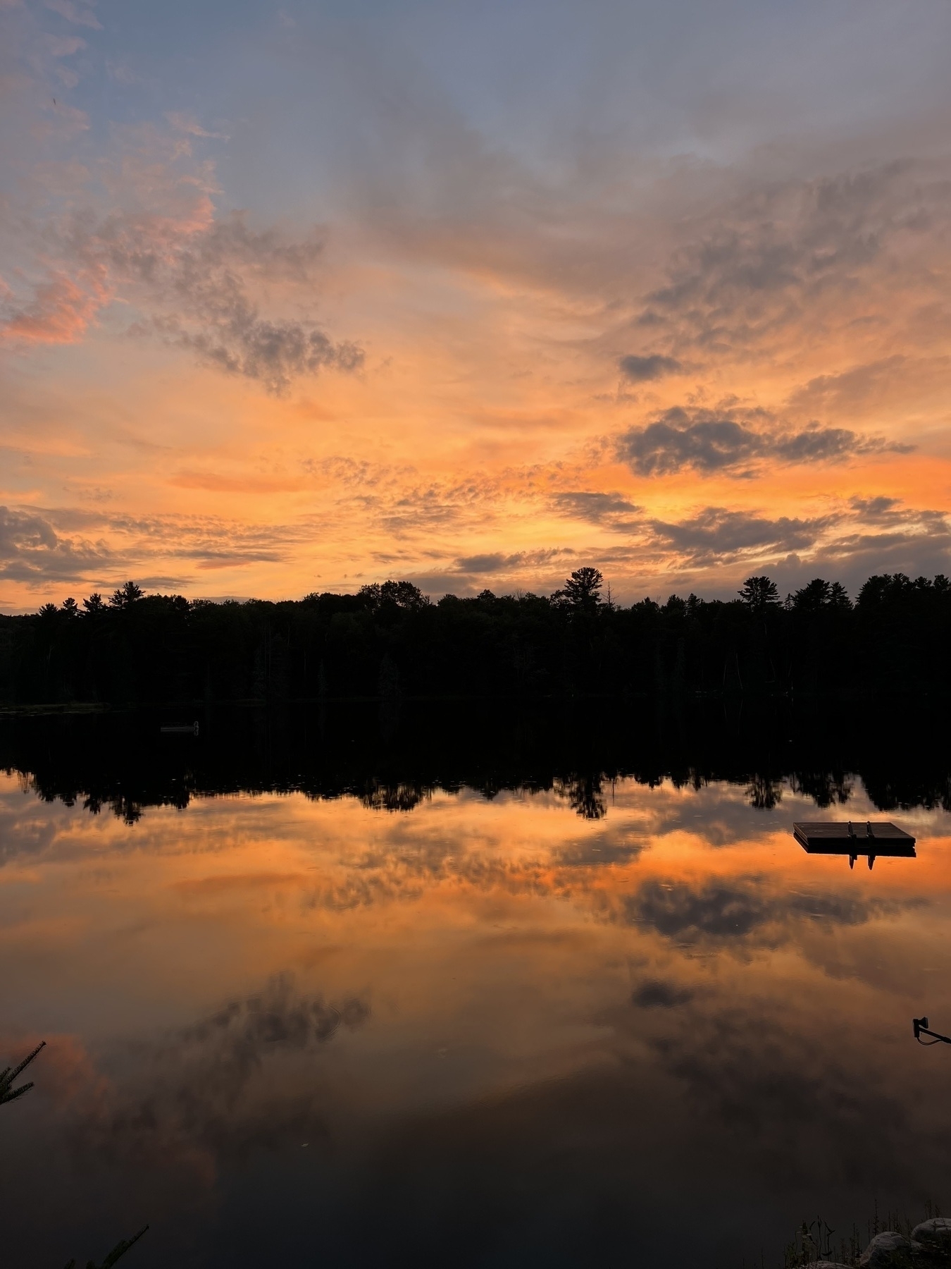 A vivid sunset with orange and blue hues reflects on a calm lake, featuring silhouetted trees and a small dock.