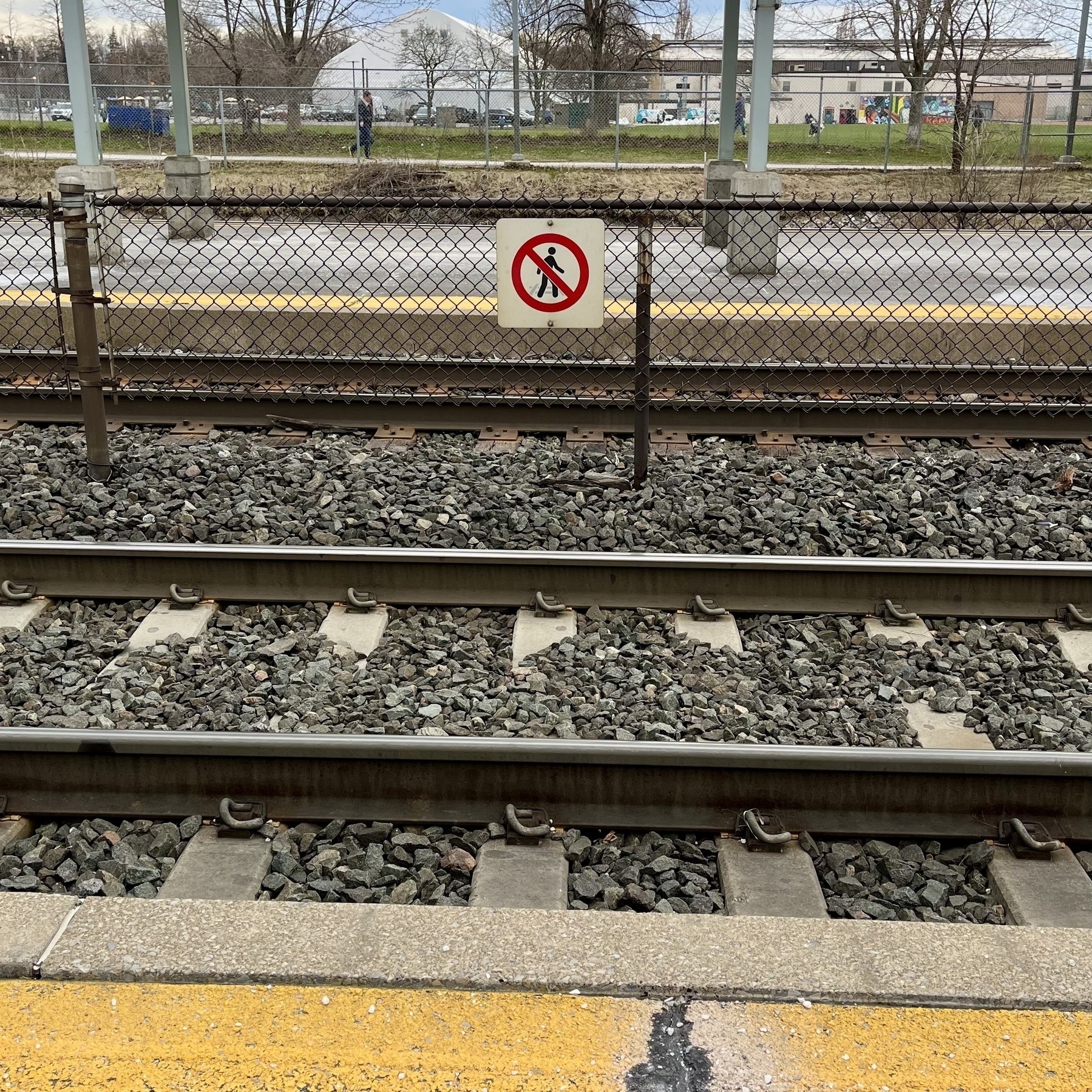 Railway tracks with a no entry sign, in front of a chain-link fence, next to a platform edge with a yellow line.