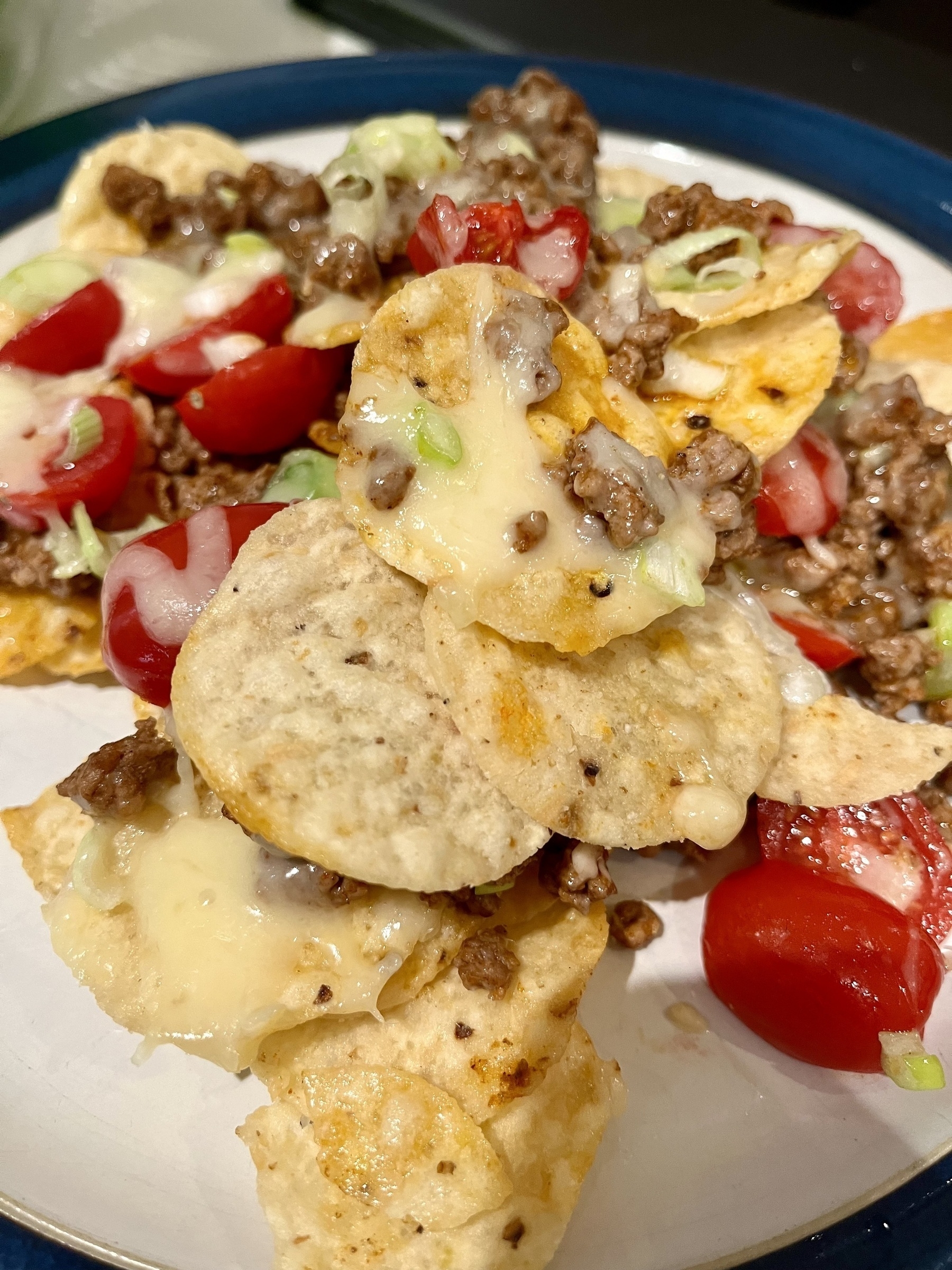 A plate of nachos topped with melted cheese, ground beef, diced tomatoes, and sliced green onions, served on a white plate with a blue rim.