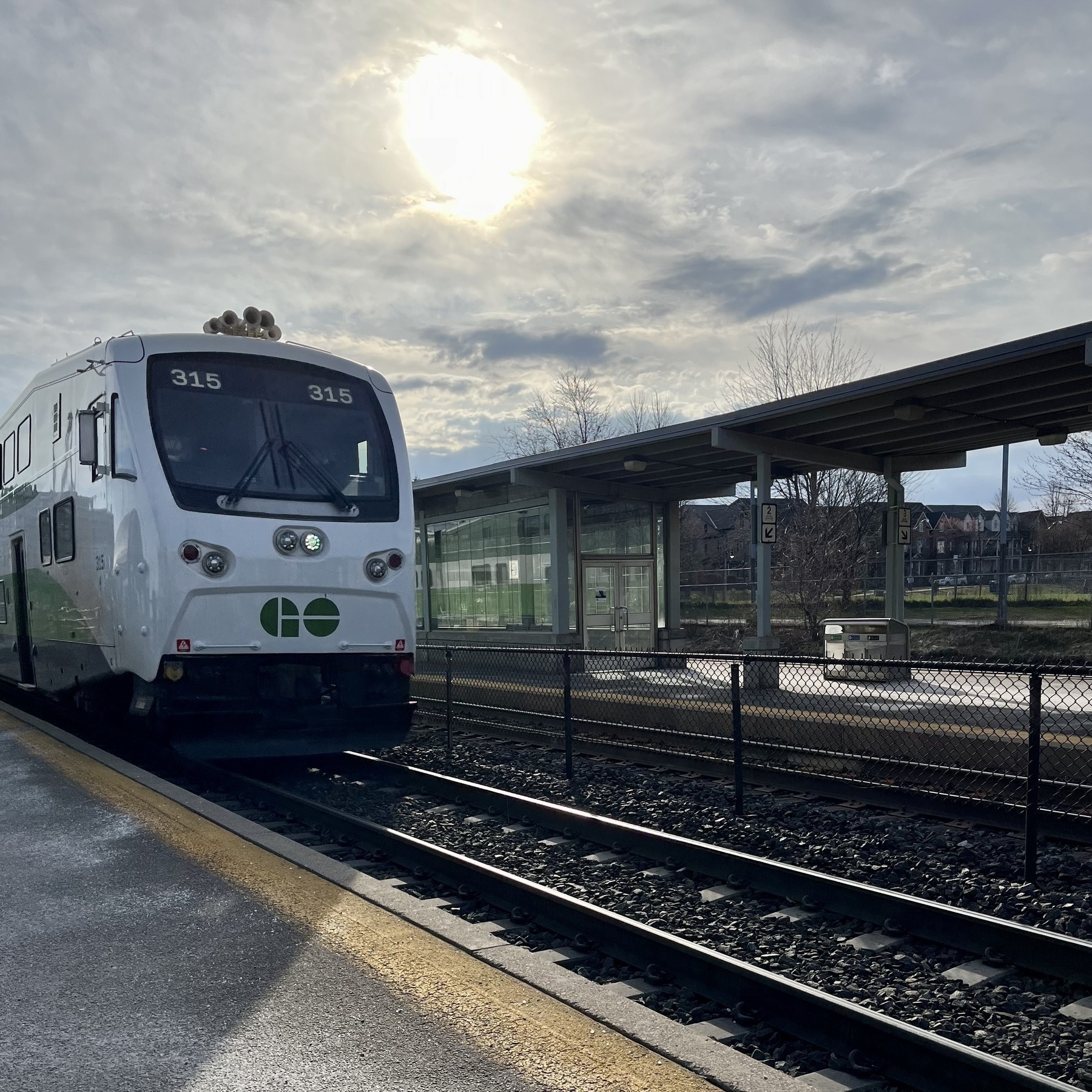 A train is stationary at a platform, with the sun partially obscured by clouds above. The setting depicts a calm, daylit train station.