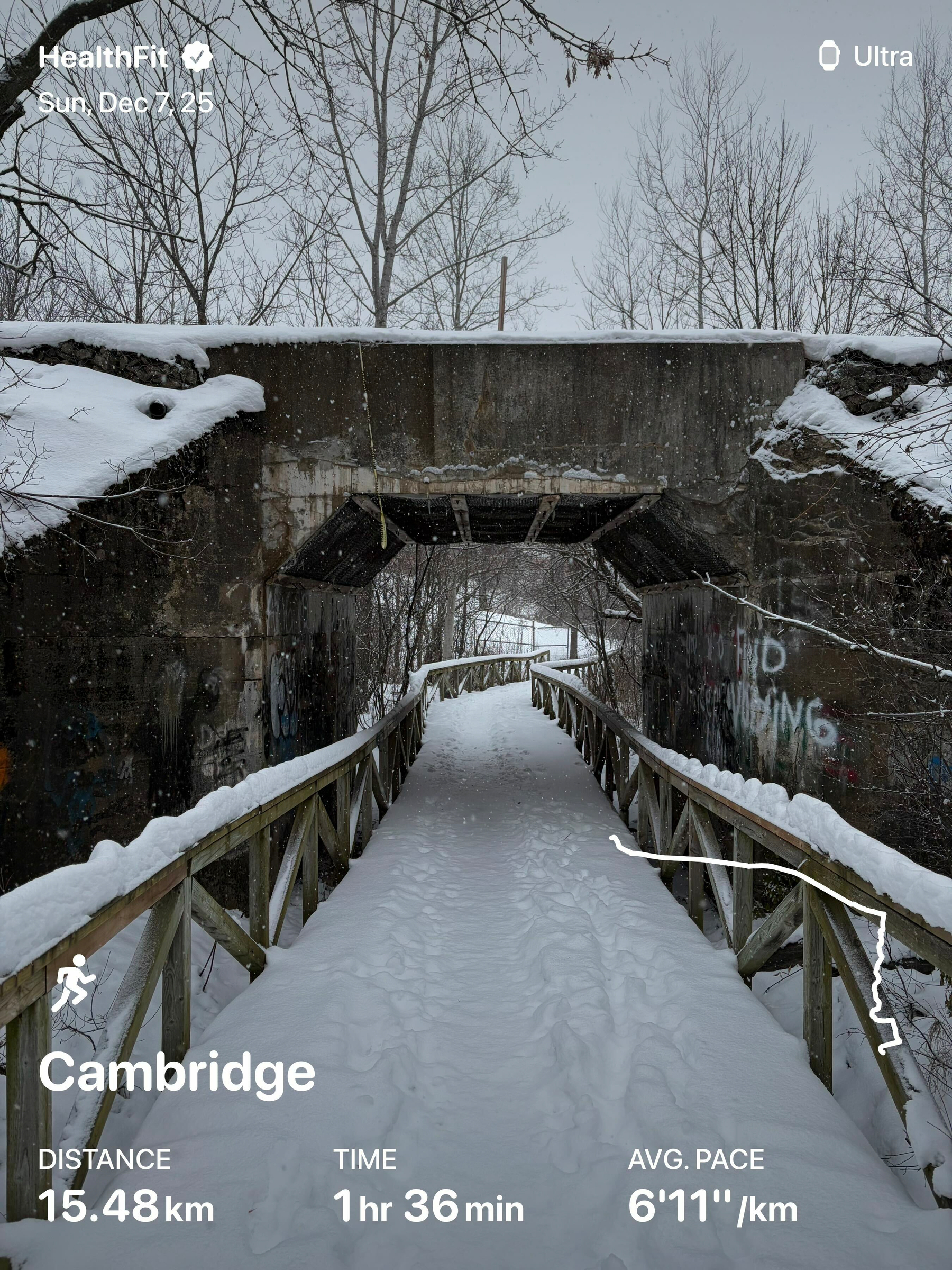 A snow-covered path leads under a graffiti-covered bridge, surrounded by bare trees, with fitness tracking information displayed at the bottom.
