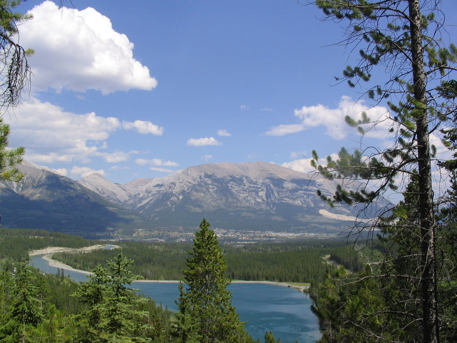 A scenic view features a clear blue lake surrounded by pine trees, with mountains in the background under a partly cloudy sky.