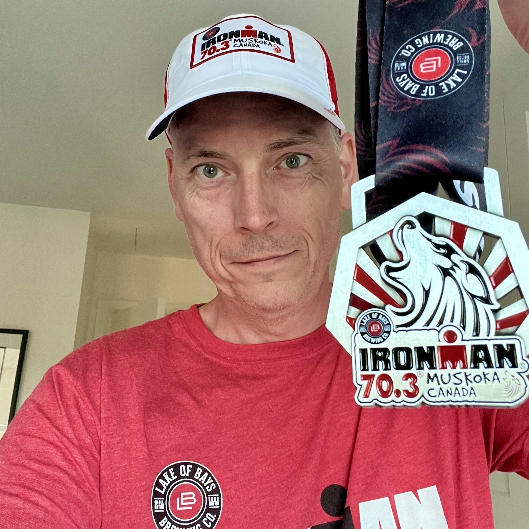 A person wearing a red shirt and white cap is holding up an Ironman Muskoka Canada medal.
