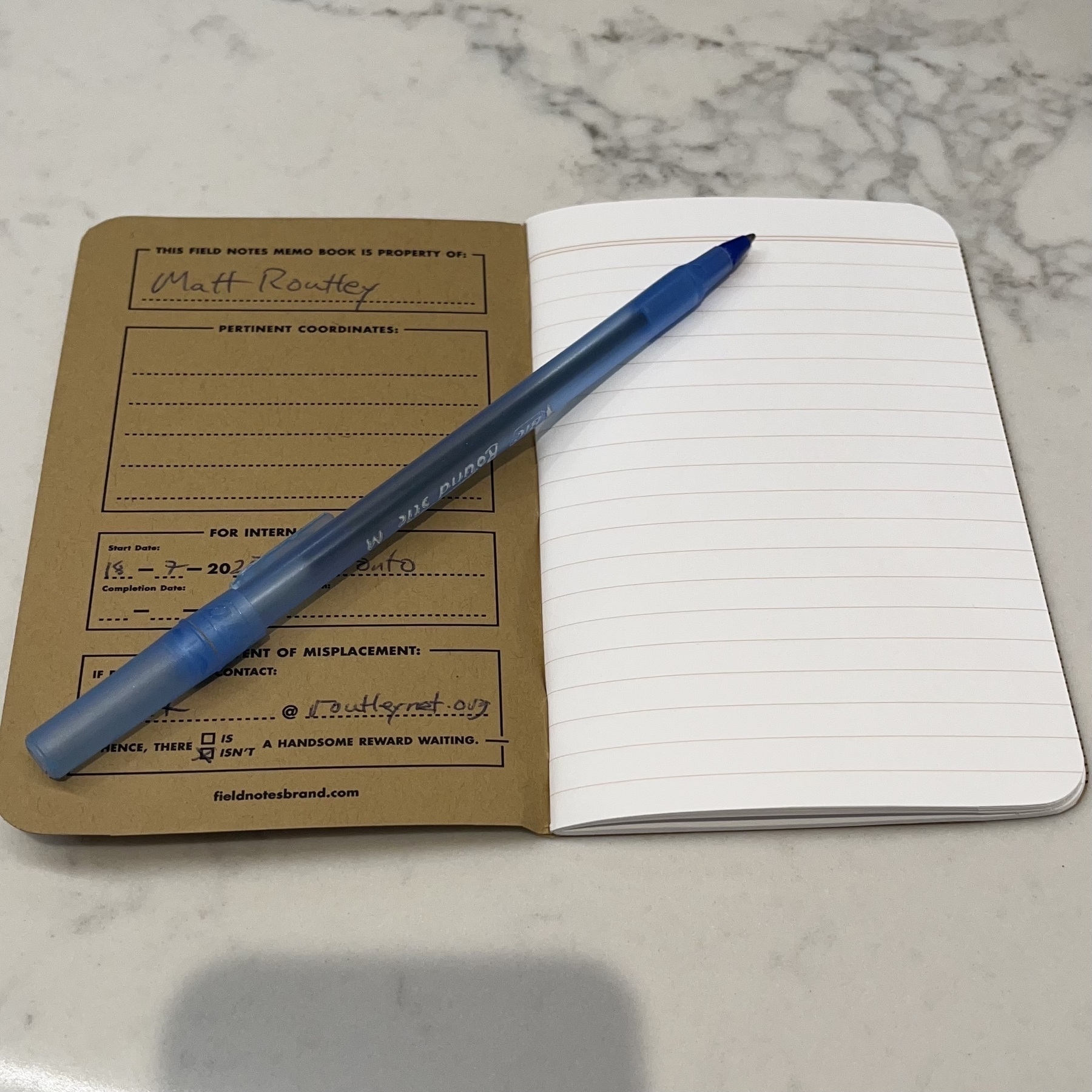 A blue pen rests on an open lined notebook placed on a marble surface.