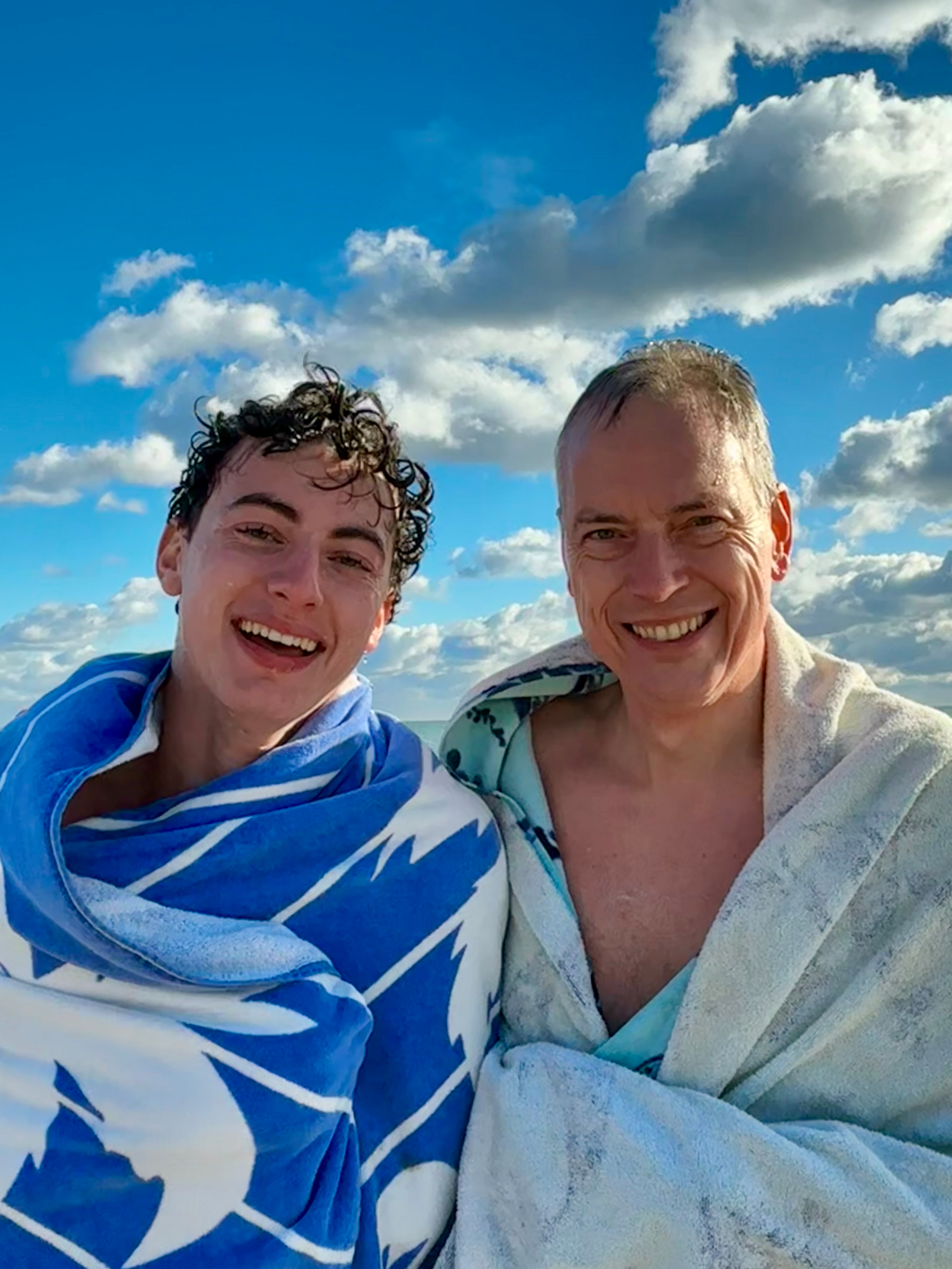 Two people are smiling while wrapped in towels against a backdrop of a blue sky with clouds.
