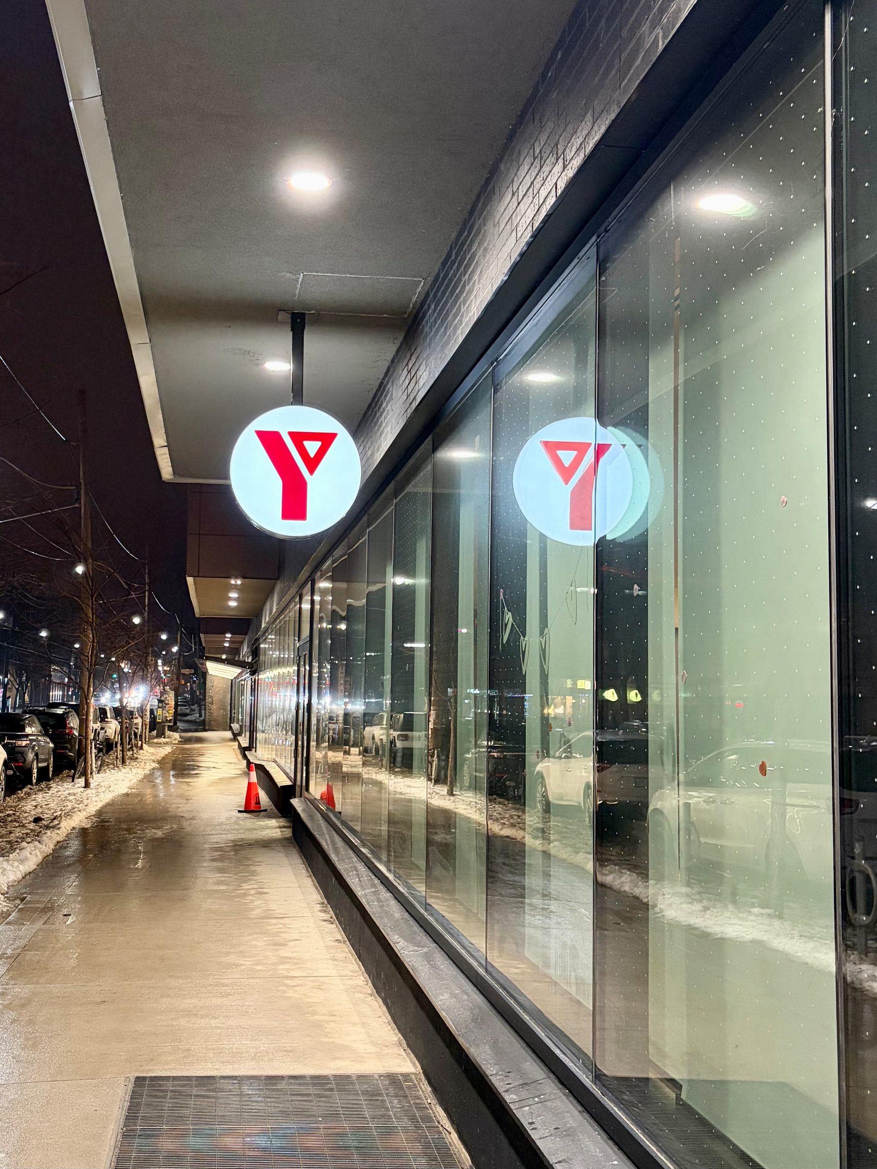 A nighttime street view features a building with large windows and illuminated red and white circular signs, reflected on the glass, along a snow-lined sidewalk.